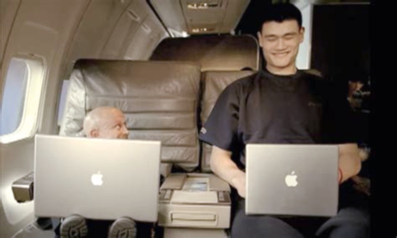 Image from an Apple commercial showing a very small Verne Troyer with a large laptop, seated on a plane next to Yao Ming with a small laptop