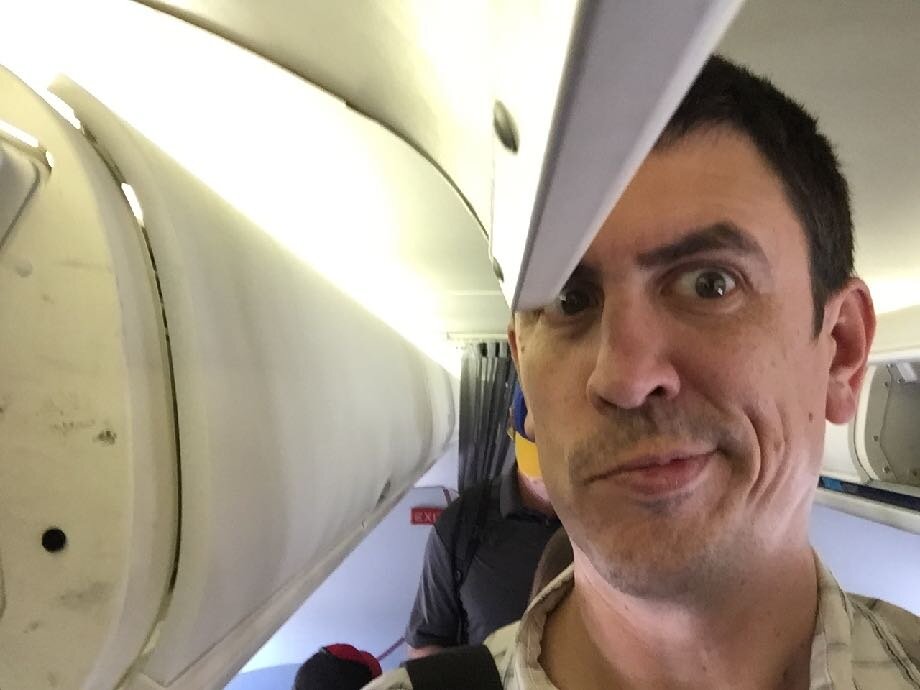 A tall man about to hit his head on the overhead bin door on an airplane