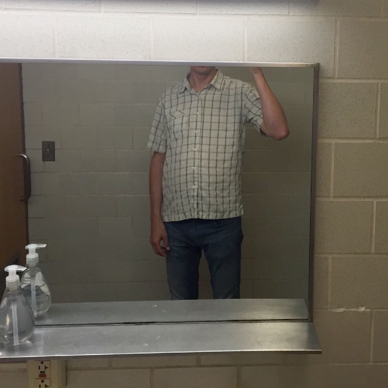 Tall man standing in front of a restaurant bathroom mirror, but only able to see himself from the neck down
